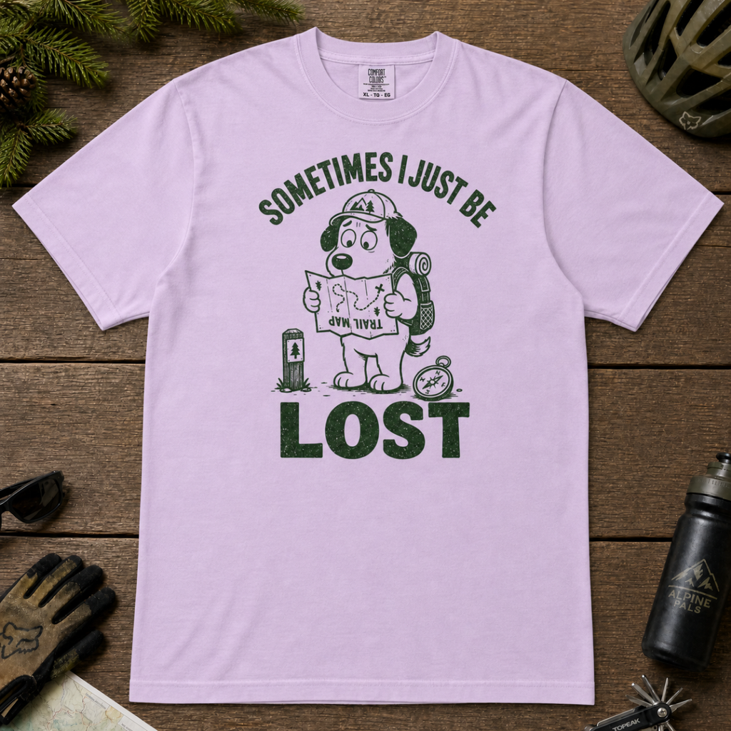 Flat lay of an orchid colored t-shirt featuring a funny lost hiker dog graphic with a trail map, compass, backpack, and bold “SOMETIMES I JUST BE LOST” text.
