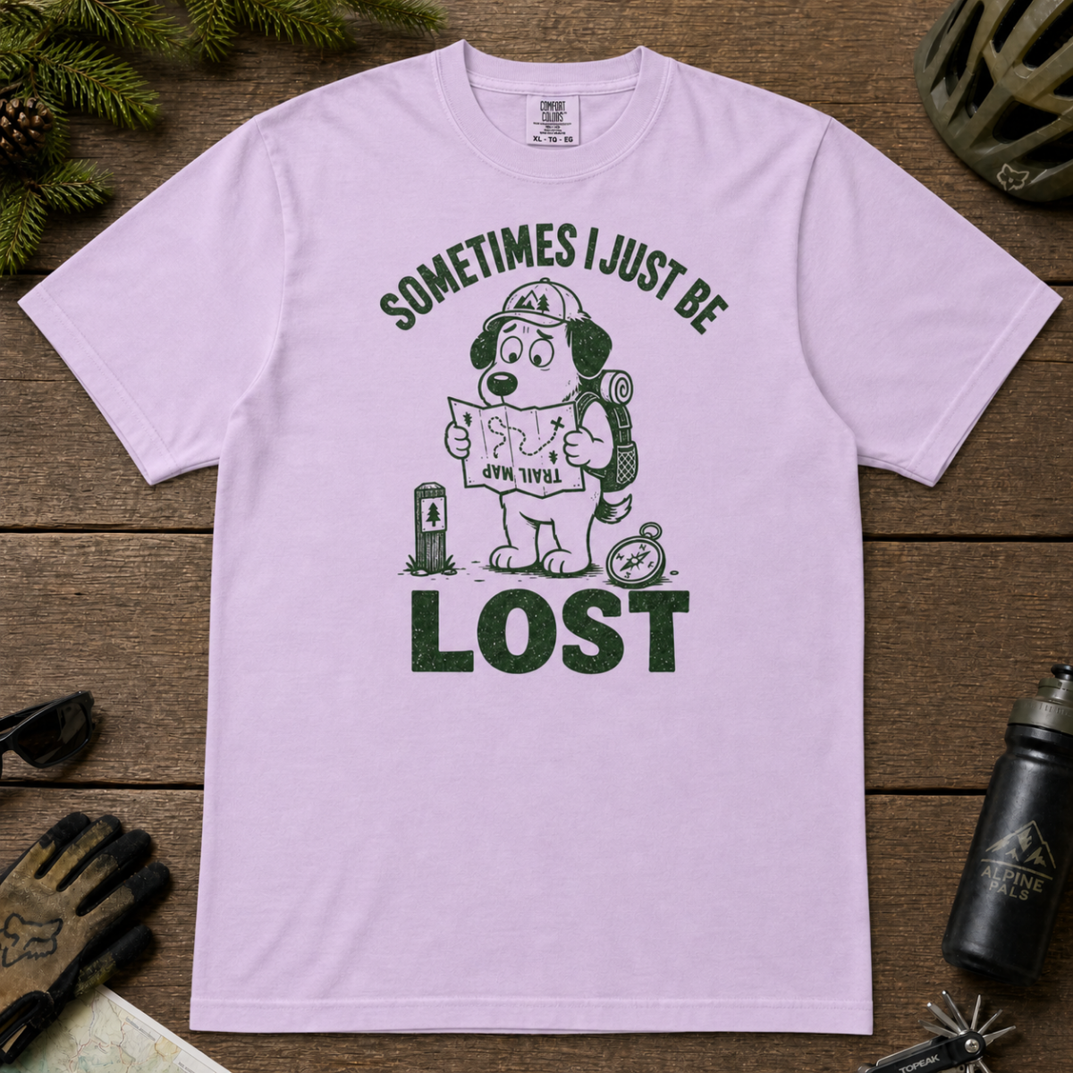 Flat lay of an orchid colored t-shirt featuring a funny lost hiker dog graphic with a trail map, compass, backpack, and bold “SOMETIMES I JUST BE LOST” text.