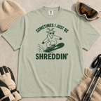 Flat lay of a bay green t-shirt featuring a green “Sometimes I Just Be Shreddin’” design with a goat snowboarding in sunglasses.