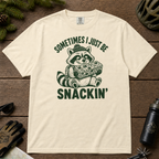 Flat lay of an ivory white t-shirt featuring a funny green “Sometimes I Just Be Snackin’” design with a happy raccoon holding a large snack bar.