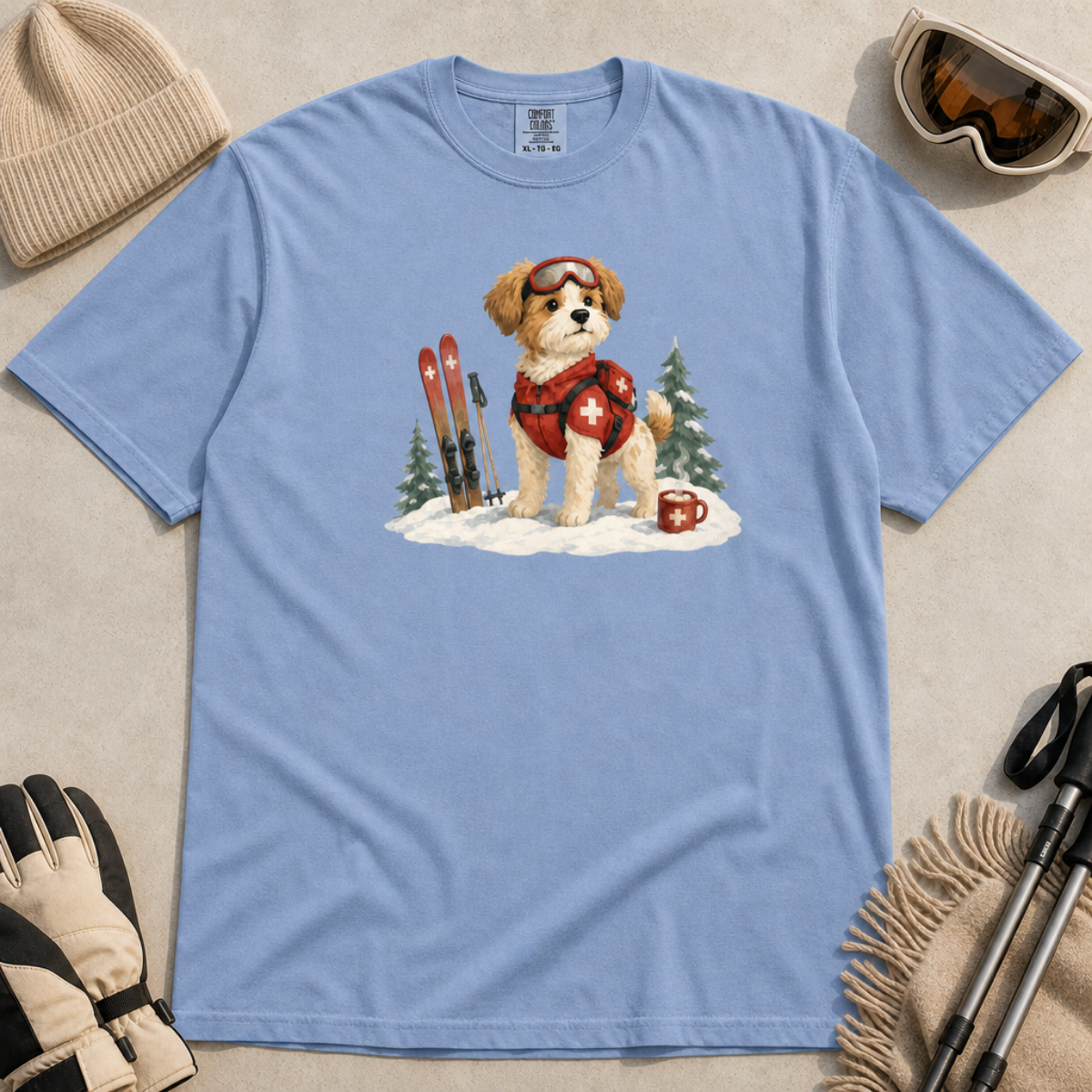 Flat lay of a washed denim blue t-shirt featuring a cute ski patrol dog design with red rescue gear, skis, snowy trees, and a hot cocoa mug.