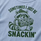 Close-up of a washed denim blue t-shirt with a playful raccoon snack graphic and bold “Sometimes I Just Be Snackin’” text.