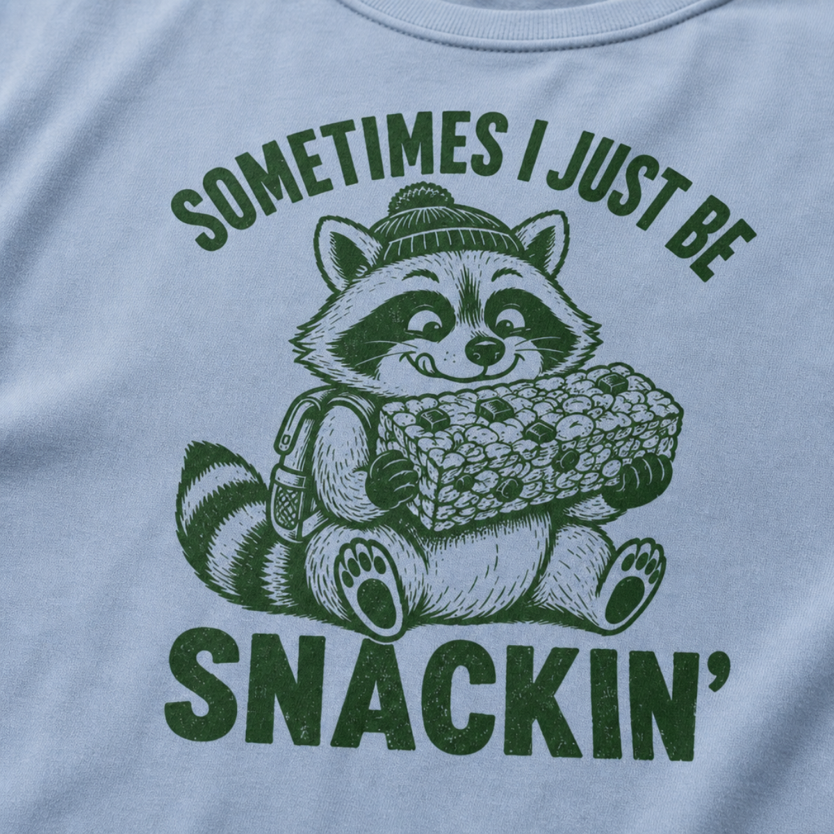 Close-up of a washed denim blue t-shirt with a playful raccoon snack graphic and bold “Sometimes I Just Be Snackin’” text.