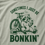Close-up of a bay green t-shirt with a funny green mountain biking design showing a tired raccoon bonking beside a bike, helmet, water bottle, and snack bar.