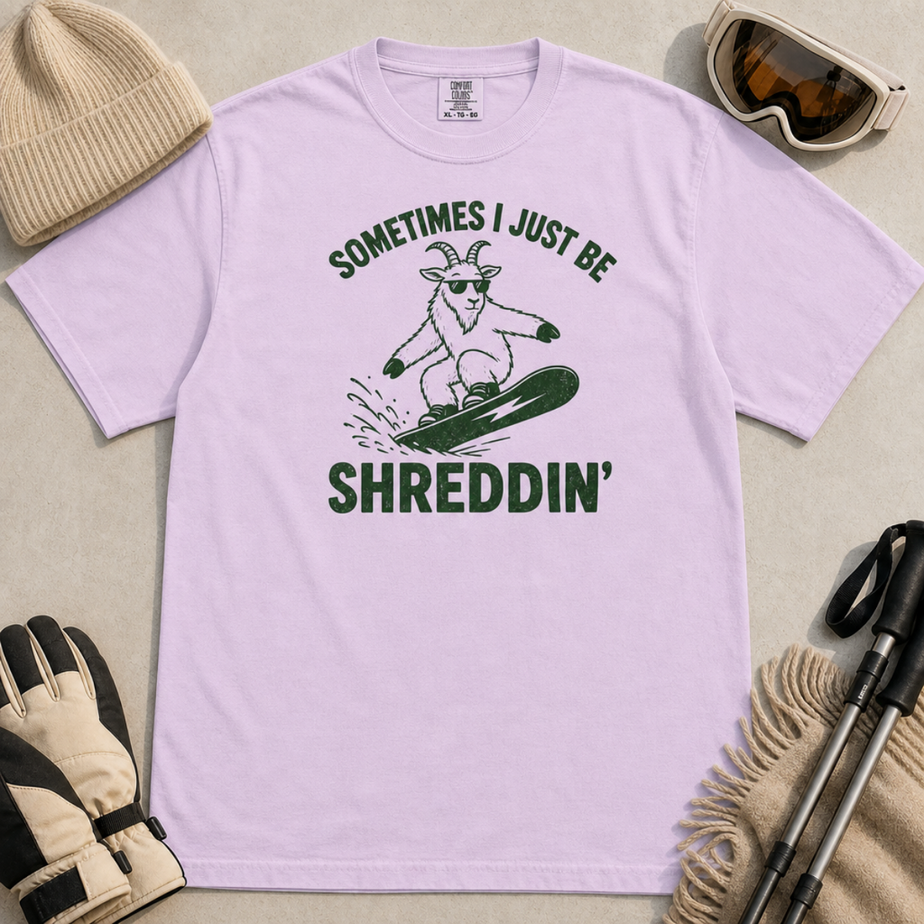 Flat lay of an orchid t-shirt featuring a green “Sometimes I Just Be Shreddin’” design with a goat snowboarding in sunglasses.