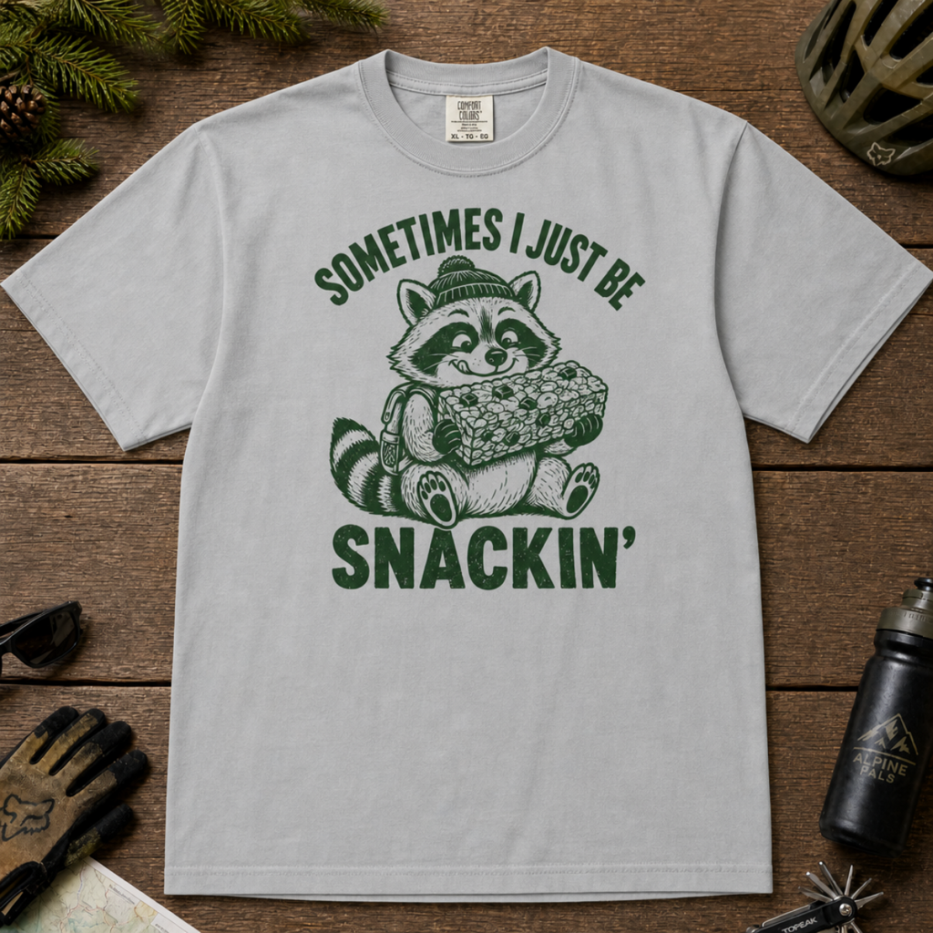 Flat lay of a granite gray t-shirt featuring a funny green “Sometimes I Just Be Snackin’” design with a happy raccoon holding a large snack bar.
