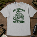 Flat lay of a granite gray t-shirt featuring a funny green “Sometimes I Just Be Snackin’” design with a happy raccoon holding a large snack bar.
