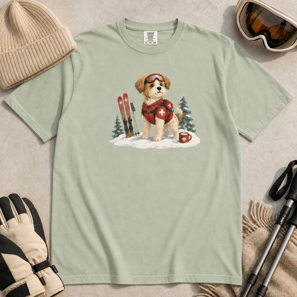 Flat lay of a bay green t-shirt featuring a cute ski patrol dog design with red rescue gear, skis, snowy trees, and a hot cocoa mug.