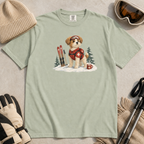 Flat lay of a bay green t-shirt featuring a cute ski patrol dog design with red rescue gear, skis, snowy trees, and a hot cocoa mug.