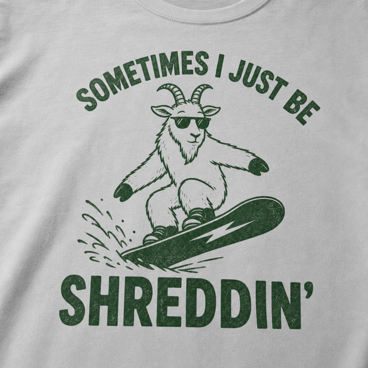 Close-up of a granite gray t-shirt with a simple green snowboard goat graphic and bold “Sometimes I Just Be Shreddin’” text.