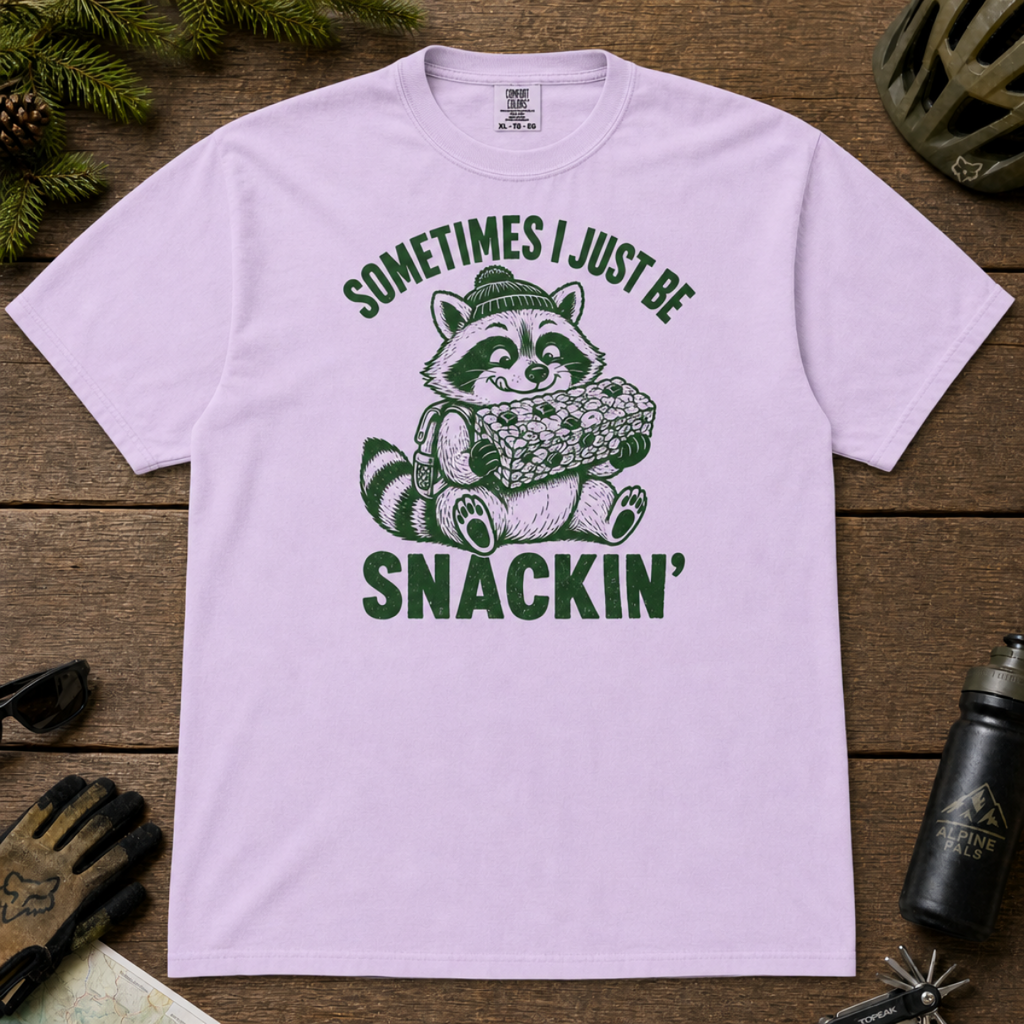 Flat lay of an orchid t-shirt featuring a funny green “Sometimes I Just Be Snackin’” design with a happy raccoon holding a large snack bar.