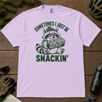 Flat lay of an orchid t-shirt featuring a funny green “Sometimes I Just Be Snackin’” design with a happy raccoon holding a large snack bar.