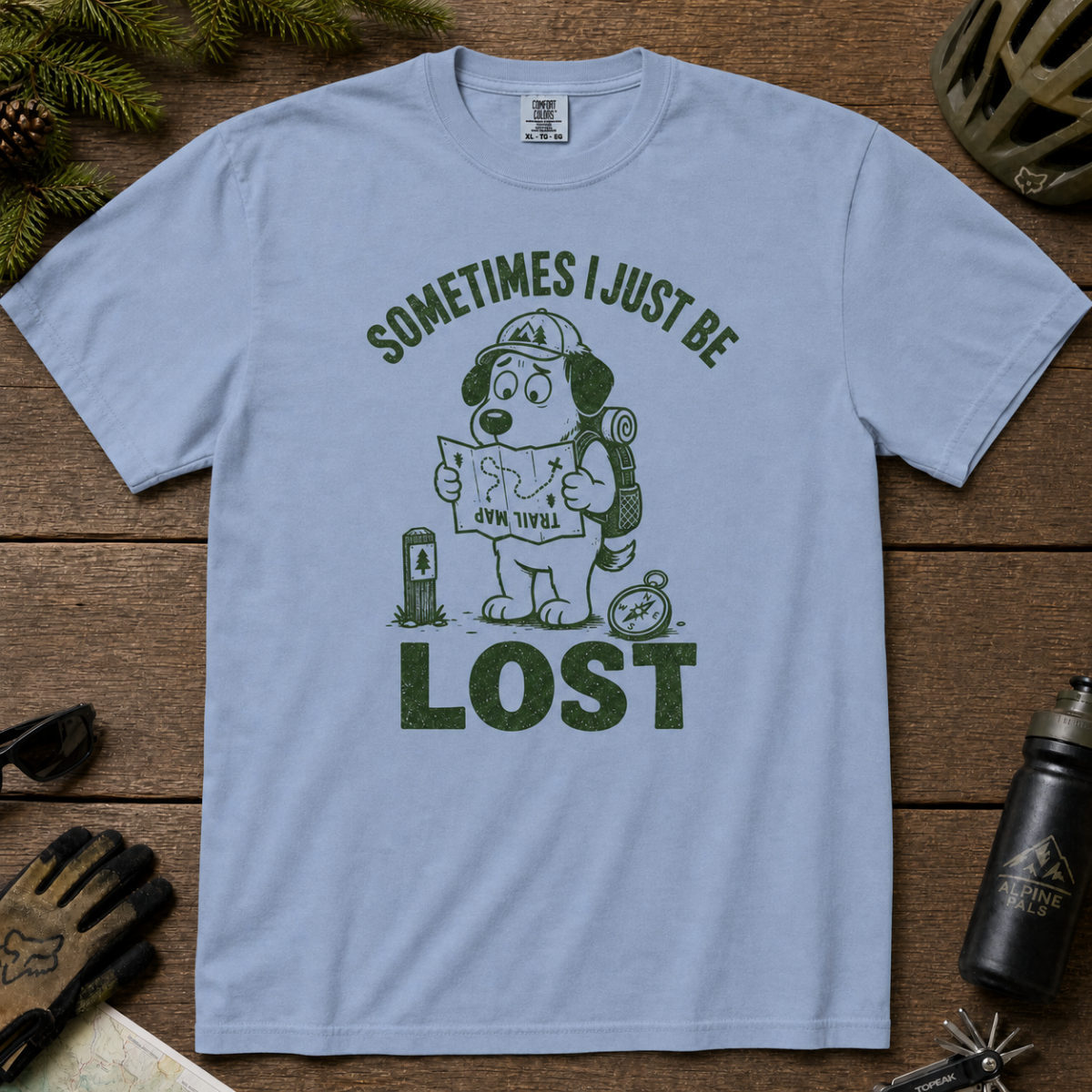 Flat lay of a washed denim blue t-shirt featuring a funny lost hiker dog graphic with a trail map, compass, backpack, and bold “SOMETIMES I JUST BE LOST” text.