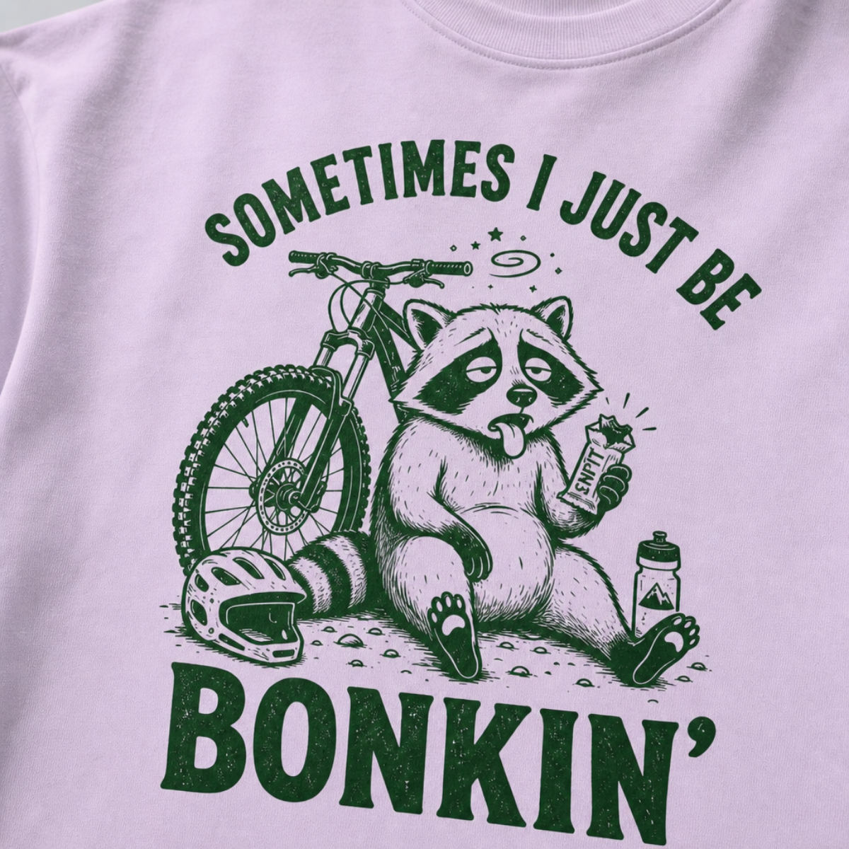 Close-up of an orchid t-shirt with a funny green mountain biking design showing a tired raccoon bonking beside a bike, helmet, water bottle, and snack bar.
