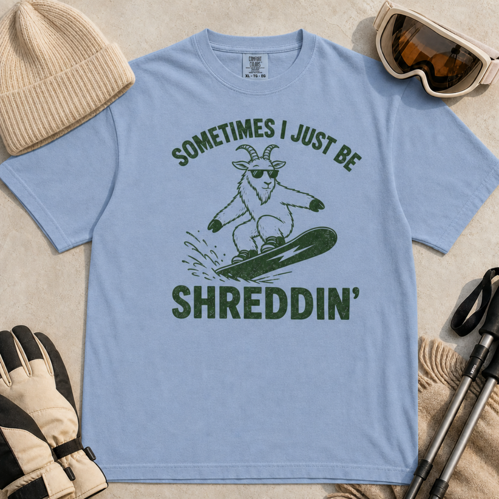 Flat lay of a white denim blue t-shirt featuring a green “Sometimes I Just Be Shreddin’” design with a goat snowboarding in sunglasses.