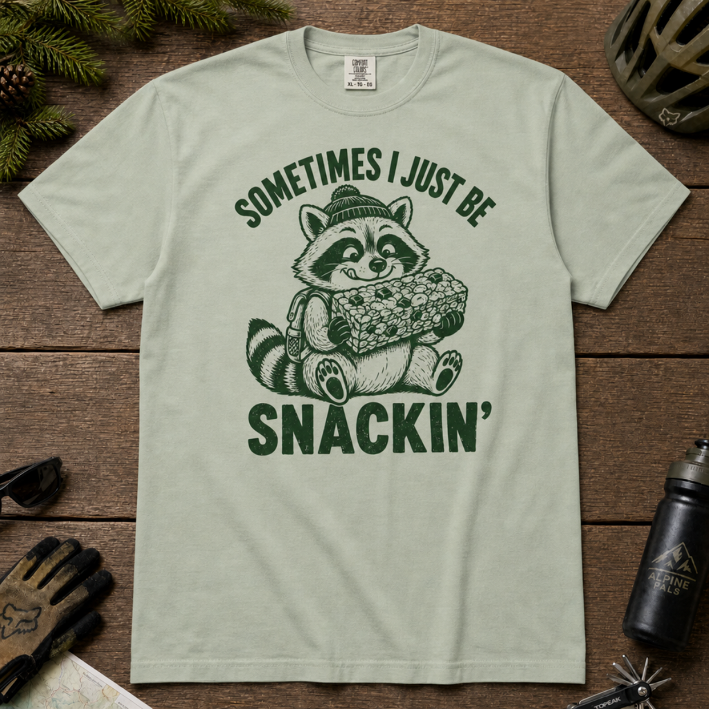 Flat lay of a bay green t-shirt featuring a funny green “Sometimes I Just Be Snackin’” design with a happy raccoon holding a large snack bar.