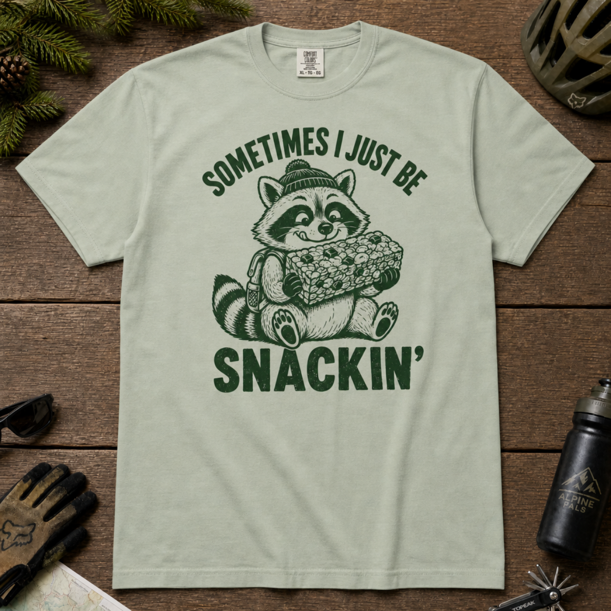 Flat lay of a bay green t-shirt featuring a funny green “Sometimes I Just Be Snackin’” design with a happy raccoon holding a large snack bar.