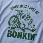 Close-up of a washed denim blue t-shirt with a funny green mountain biking design showing a tired raccoon bonking beside a bike, helmet, water bottle, and snack bar.
