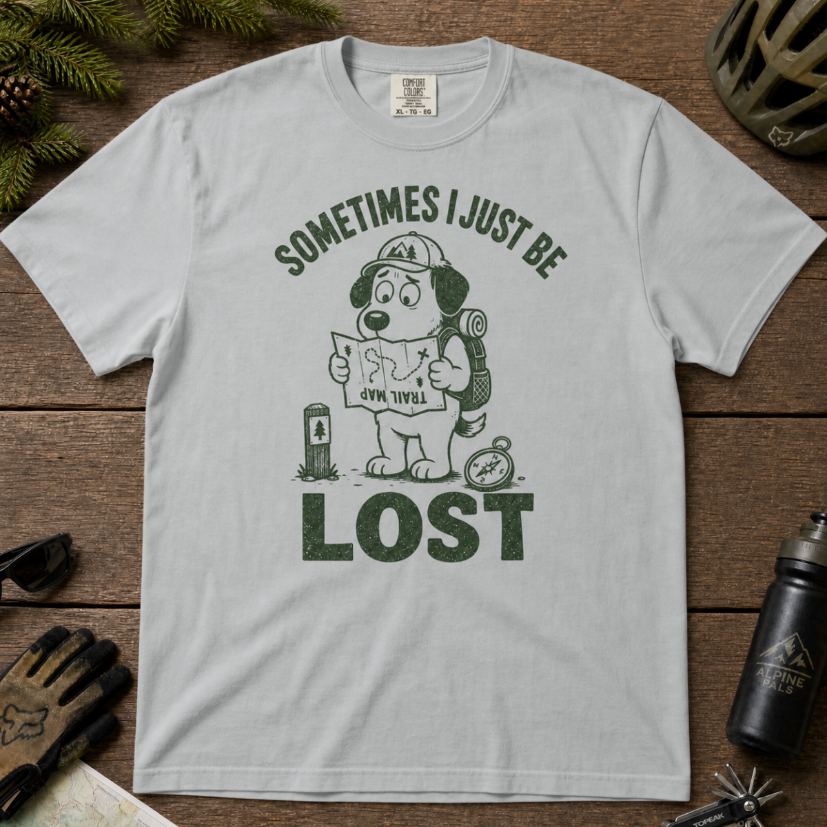 Flat lay of a granite gray t-shirt featuring a funny lost hiker dog graphic with a trail map, compass, backpack, and bold “SOMETIMES I JUST BE LOST” text.