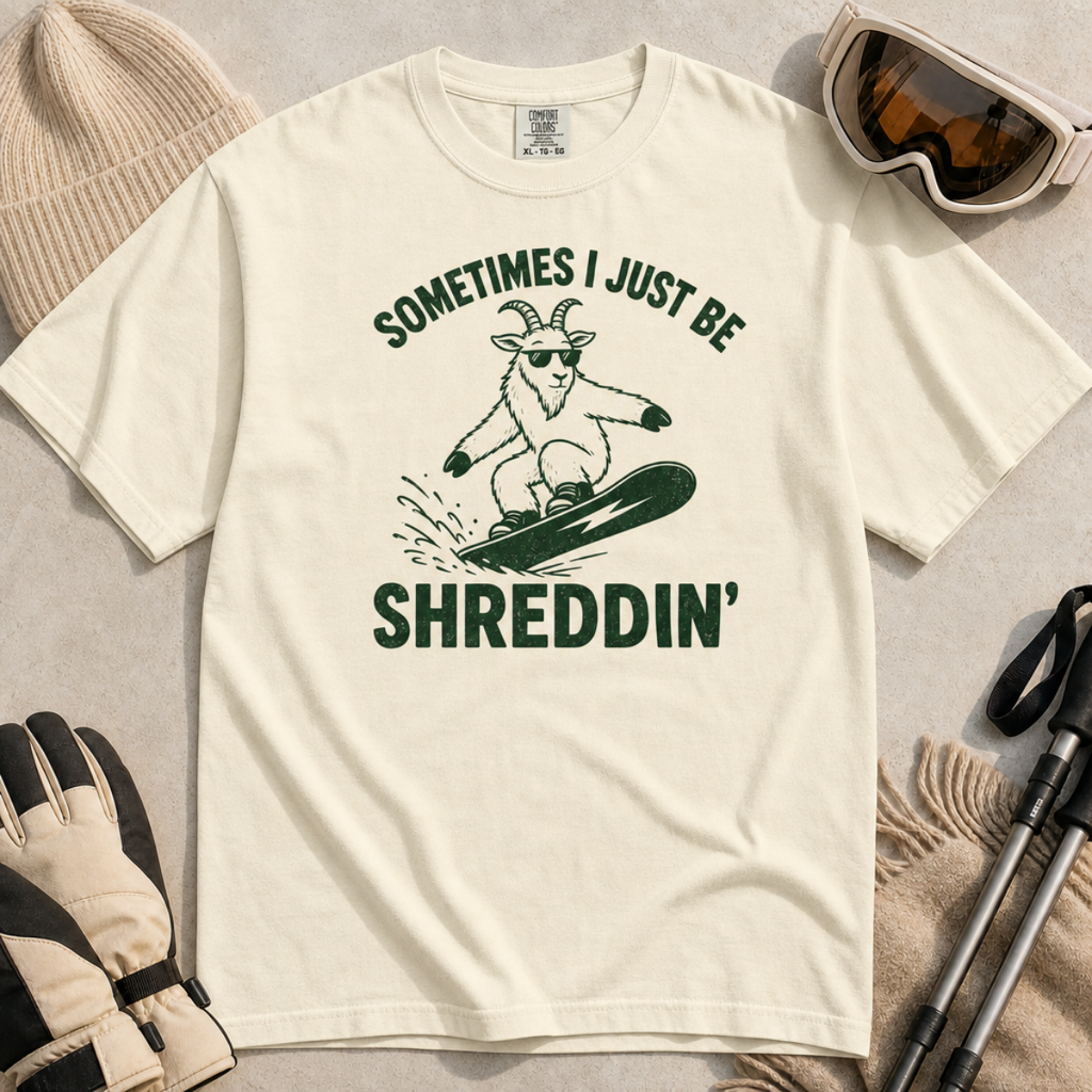 Flat lay of an ivory white t-shirt featuring a green “Sometimes I Just Be Shreddin’” design with a goat snowboarding in sunglasses.