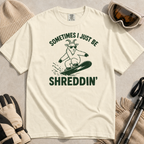 Flat lay of an ivory white t-shirt featuring a green “Sometimes I Just Be Shreddin’” design with a goat snowboarding in sunglasses.