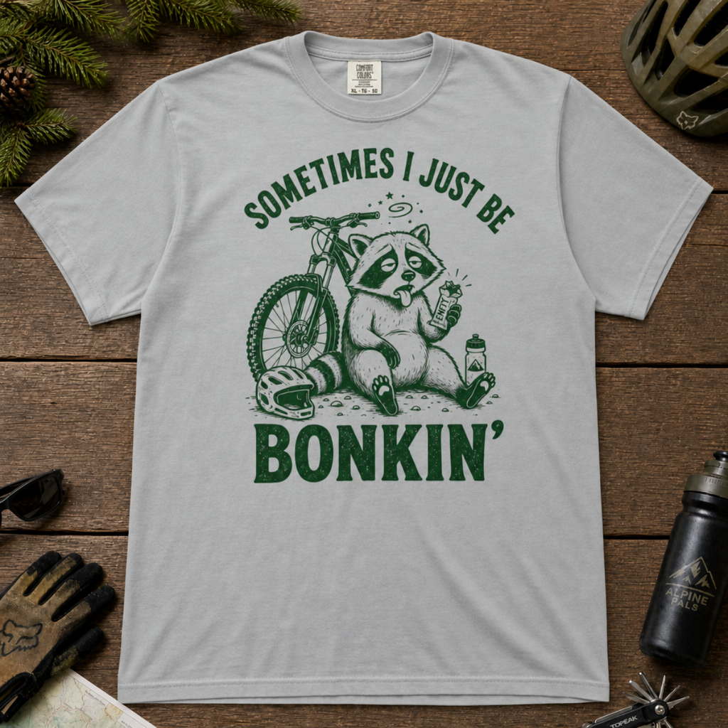 Flat lay of a granite gray t-shirt featuring a “Sometimes I Just Be Bonkin’” raccoon mountain biking graphic, styled with trail accessories on a wooden background.
