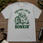 Flat lay of a granite gray t-shirt featuring a “Sometimes I Just Be Bonkin’” raccoon mountain biking graphic, styled with trail accessories on a wooden background.