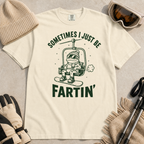 Flat lay of an ivory white t-shirt featuring a funny green “Sometimes I Just Be Fartin’” design with a frog riding a chairlift on a snowboard.