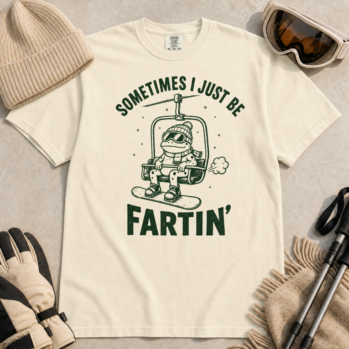 Flat lay of an ivory white t-shirt featuring a funny green “Sometimes I Just Be Fartin’” design with a frog riding a chairlift on a snowboard.