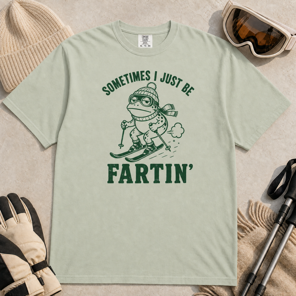 Flat lay of a bay green t-shirt featuring a funny frog skiing graphic with “Sometimes I Just Be Fartin’” text, styled with winter accessories.