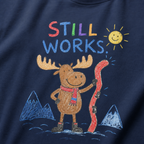 Close-up of a navy t-shirt with a playful crayon-style ski design showing a smiling moose holding a red ski, with “Still Works.” text.