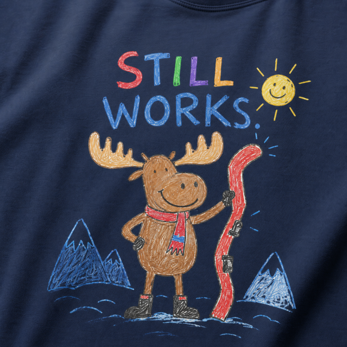Close-up of a navy t-shirt with a playful crayon-style ski design showing a smiling moose holding a red ski, with “Still Works.” text.