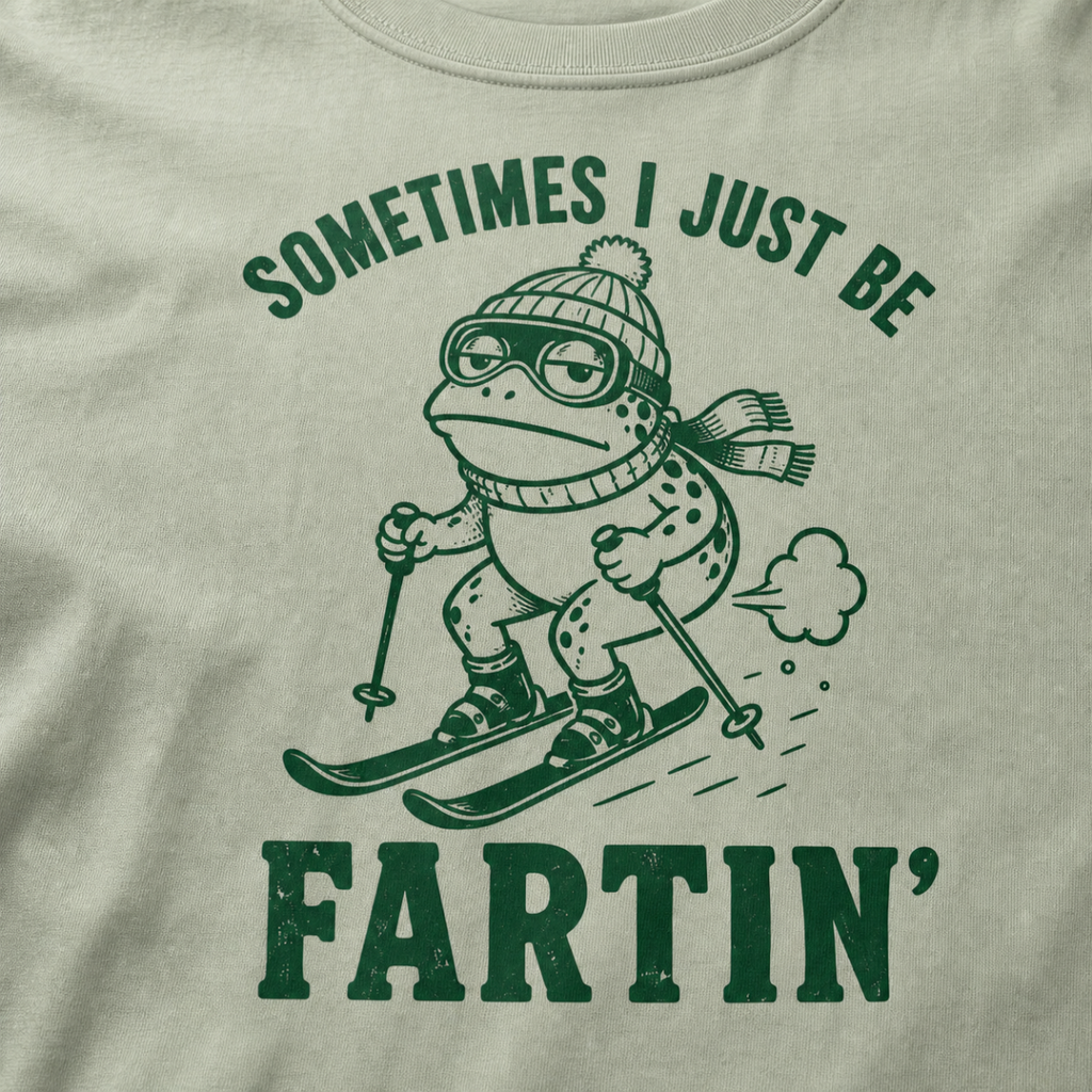 Close-up of a bay green t-shirt with a green “Sometimes I Just Be Fartin’” ski design showing a deadpan frog skiing with a small puff behind it.