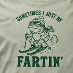 Close-up of a bay green t-shirt with a green “Sometimes I Just Be Fartin’” ski design showing a deadpan frog skiing with a small puff behind it.