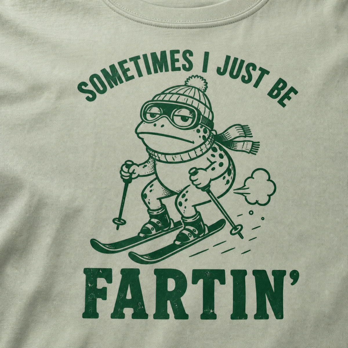 Close-up of a bay green t-shirt with a green “Sometimes I Just Be Fartin’” ski design showing a deadpan frog skiing with a small puff behind it.