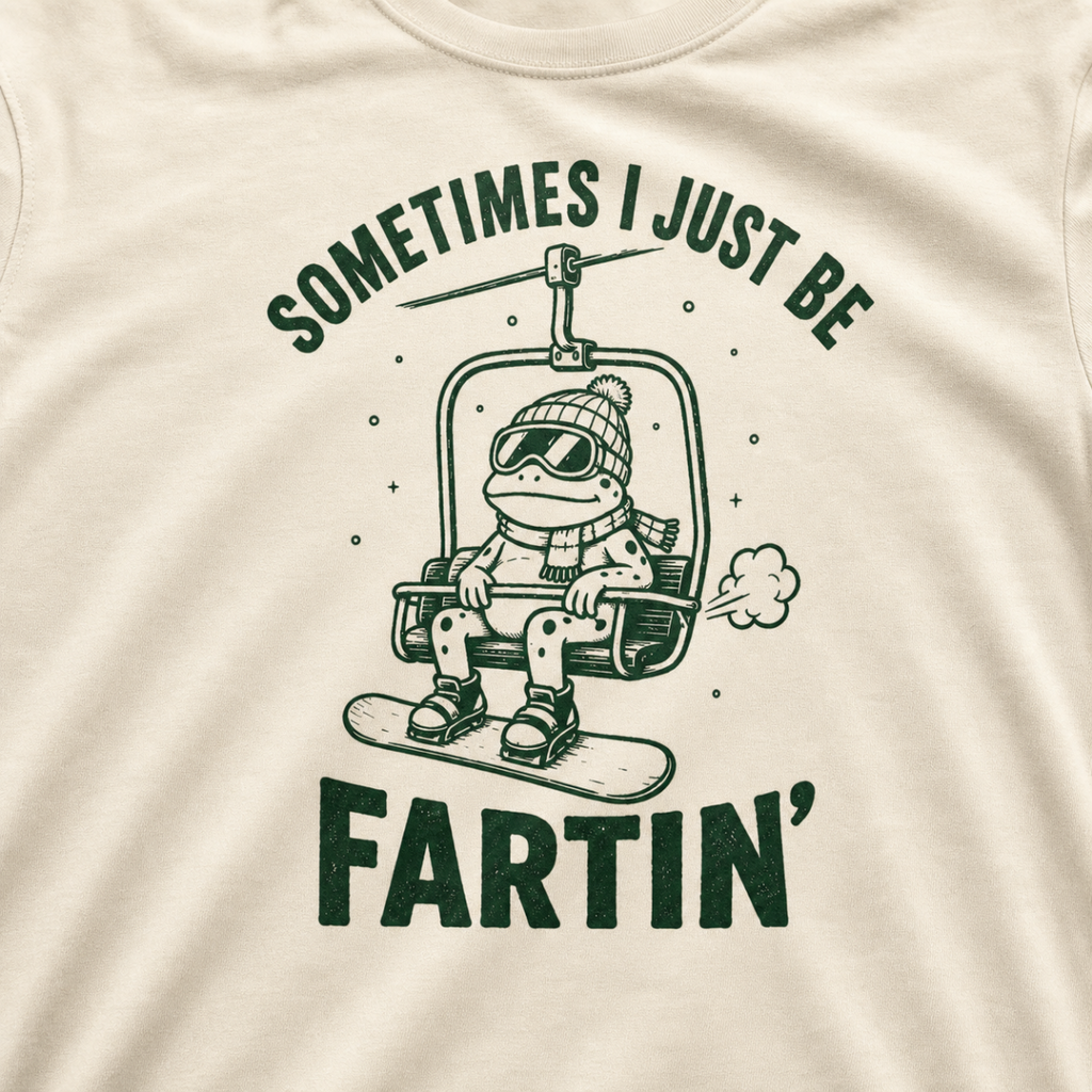 Close-up of an ivory white t-shirt with a playful frog chairlift graphic and bold “Sometimes I Just Be Fartin’” text.