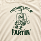 Close-up of an ivory white t-shirt with a playful frog chairlift graphic and bold “Sometimes I Just Be Fartin’” text.