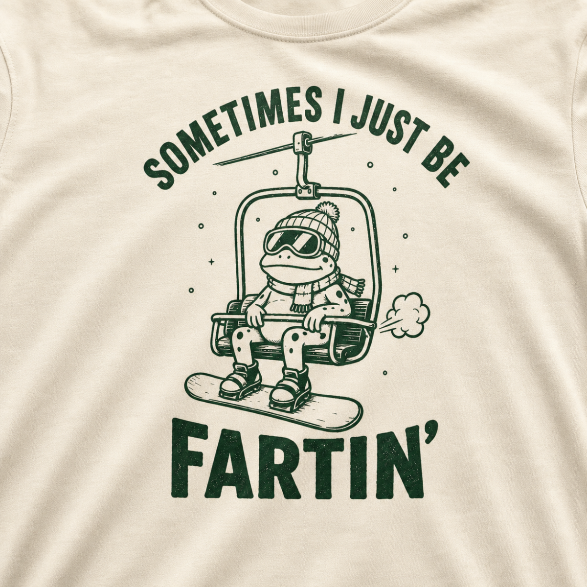 Close-up of an ivory white t-shirt with a playful frog chairlift graphic and bold “Sometimes I Just Be Fartin’” text.
