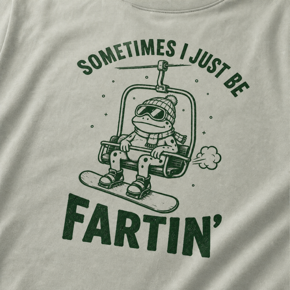 Close-up of a bay green t-shirt with a playful frog chairlift graphic and bold “Sometimes I Just Be Fartin’” text.