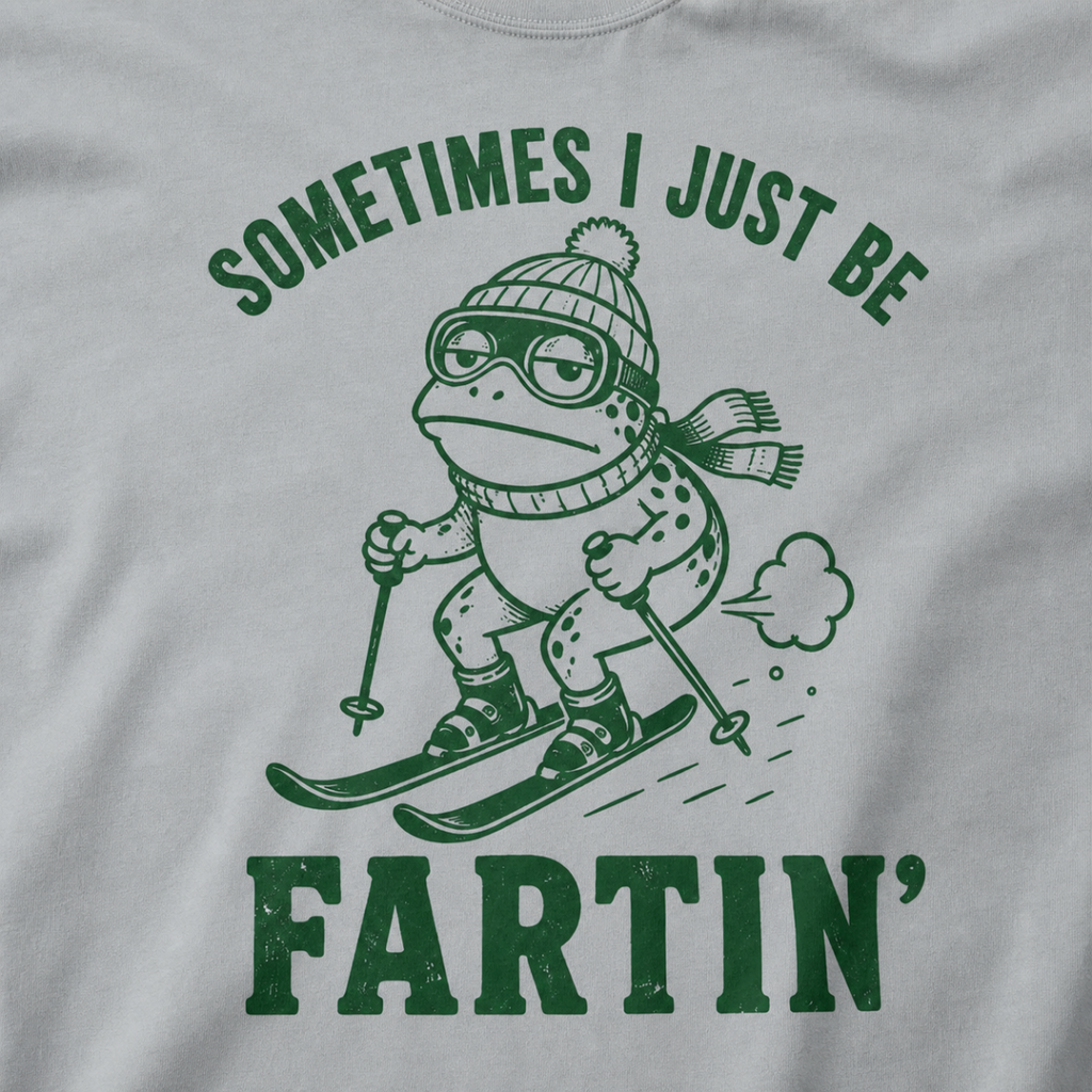 Close-up of a granite gray t-shirt with a green “Sometimes I Just Be Fartin’” ski design showing a deadpan frog skiing with a small puff behind it.