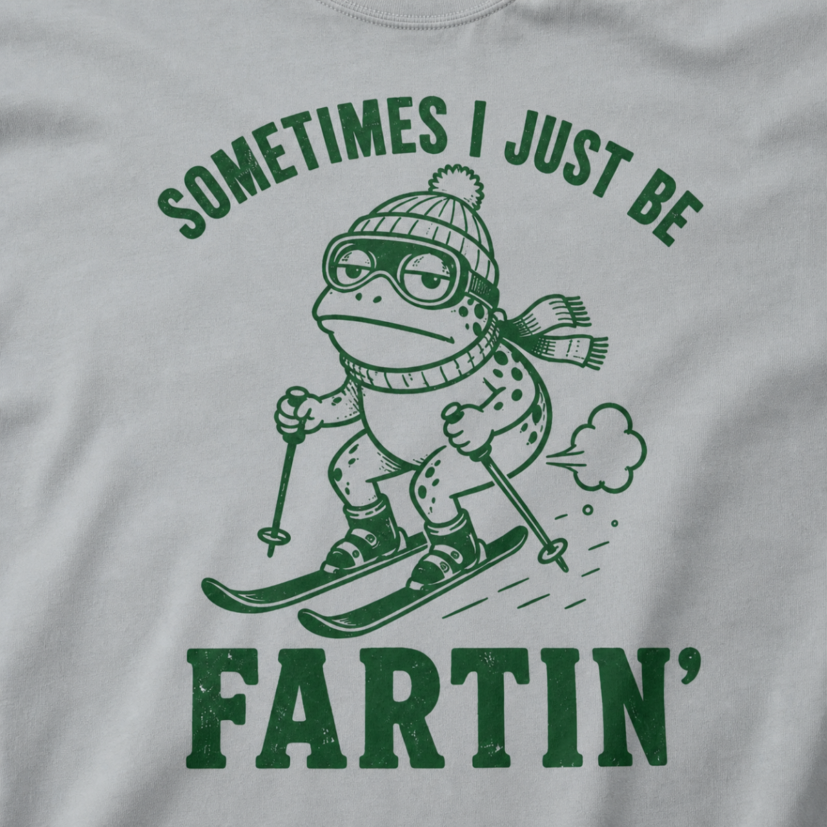 Close-up of a granite gray t-shirt with a green “Sometimes I Just Be Fartin’” ski design showing a deadpan frog skiing with a small puff behind it.