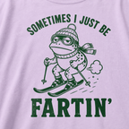 Close-up of an orchid colored t-shirt with a green “Sometimes I Just Be Fartin’” ski design showing a deadpan frog skiing with a small puff behind it.