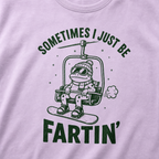 Close-up of an orchid colored t-shirt with a playful frog chairlift graphic and bold “Sometimes I Just Be Fartin’” text.
