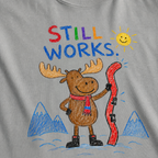 Close-up of a granite gray t-shirt with a playful crayon-style ski design showing a smiling moose holding a red ski, with “Still Works.” text.