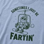 Close-up of a washed denim blue t-shirt with a playful frog chairlift graphic and bold “Sometimes I Just Be Fartin’” text.