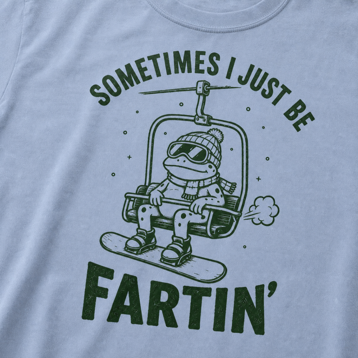 Close-up of a washed denim blue t-shirt with a playful frog chairlift graphic and bold “Sometimes I Just Be Fartin’” text.