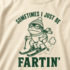 Close-up of an ivory white t-shirt with a green “Sometimes I Just Be Fartin’” ski design showing a deadpan frog skiing with a small puff behind it.