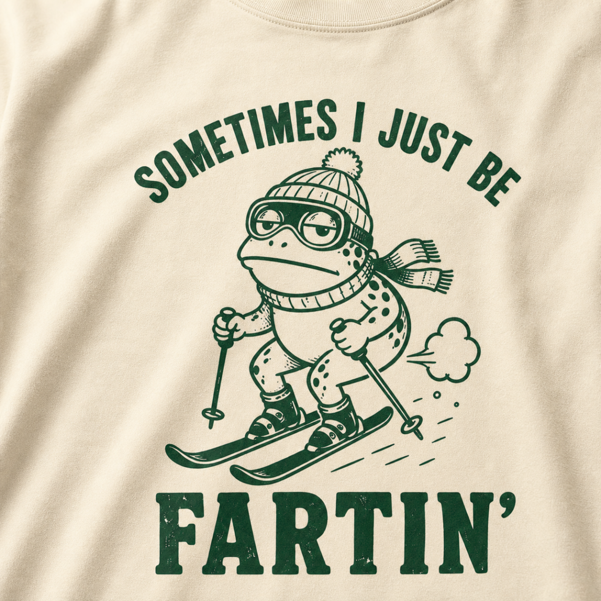 Close-up of an ivory white t-shirt with a green “Sometimes I Just Be Fartin’” ski design showing a deadpan frog skiing with a small puff behind it.