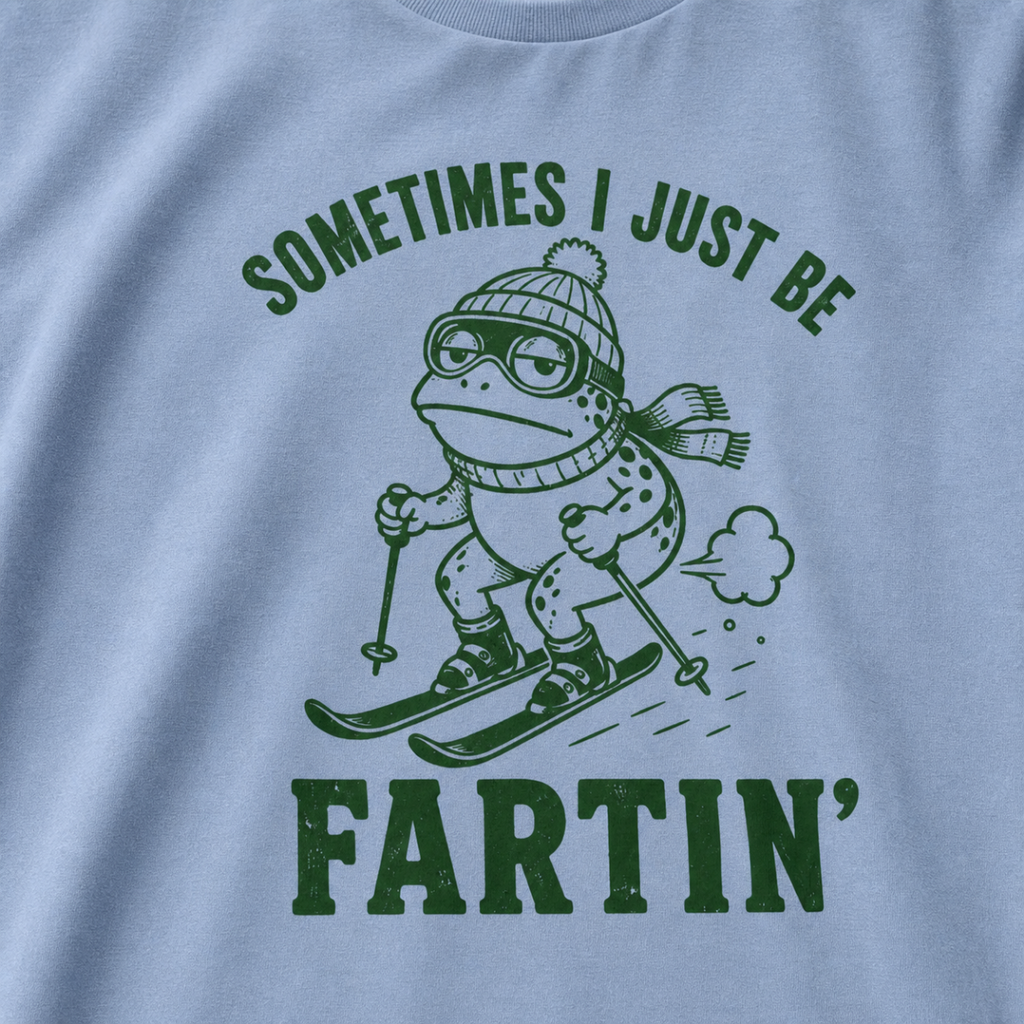 Close-up of a washed denim blue t-shirt with a green “Sometimes I Just Be Fartin’” ski design showing a deadpan frog skiing with a small puff behind it.
