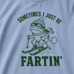 Close-up of a washed denim blue t-shirt with a green “Sometimes I Just Be Fartin’” ski design showing a deadpan frog skiing with a small puff behind it.